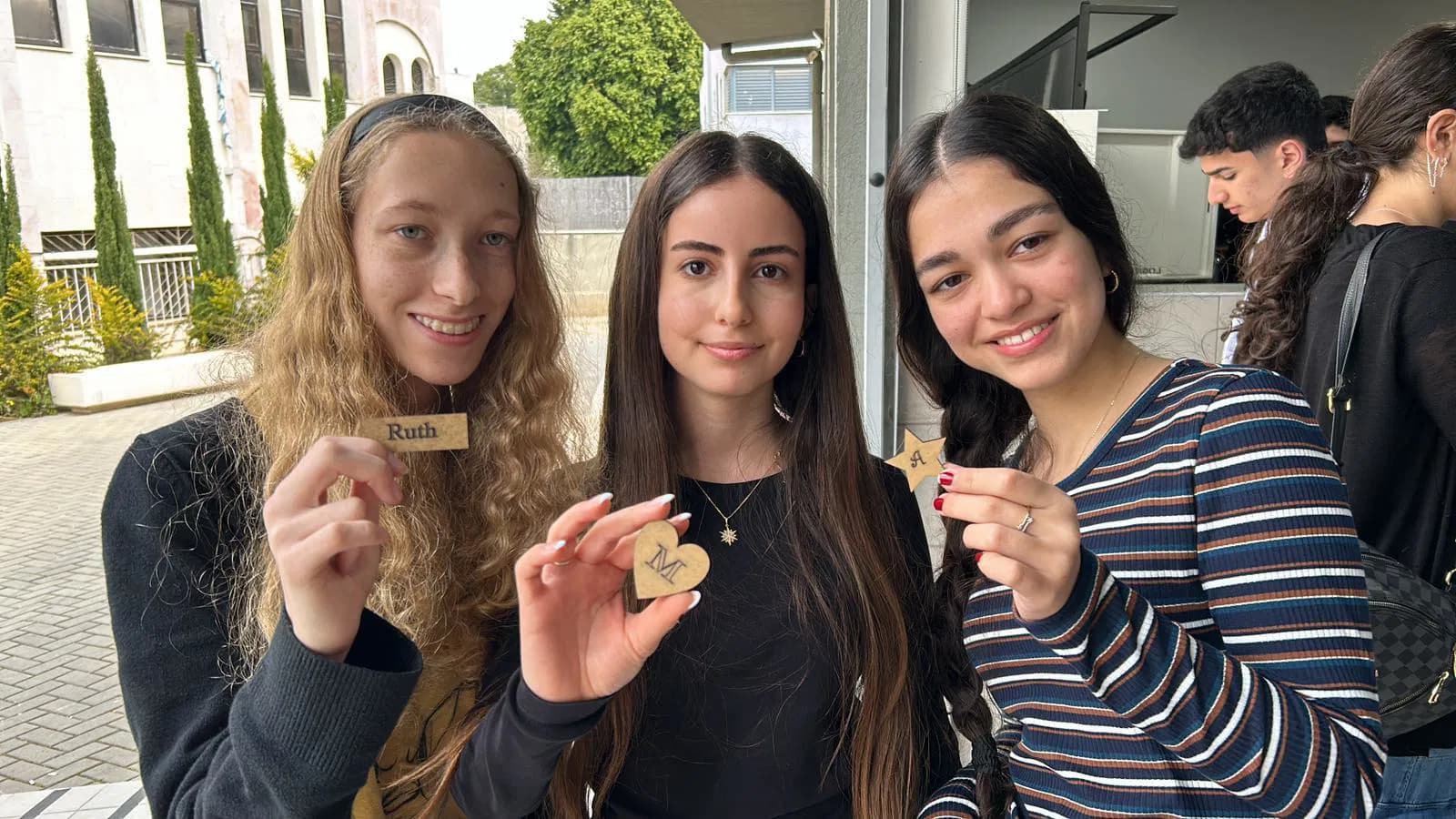 Three smiling young women hold small wooden tokens shaped like a rectangle, heart, and star.